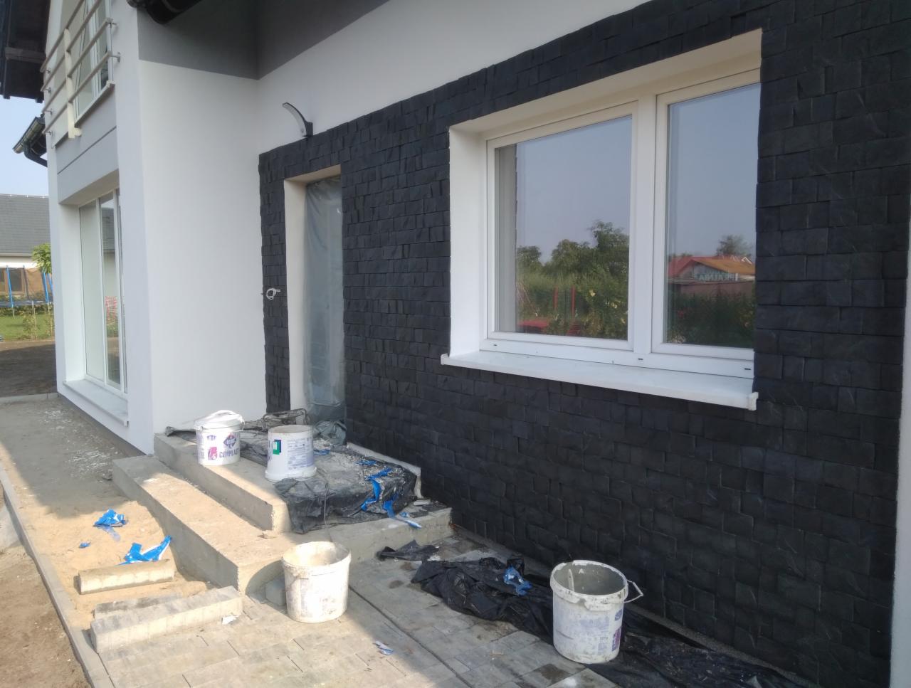 Fragment elewacji domu z czarną, kamienną okładziną, białe okna, widoczne wiadra z farbą i zabezpieczenia na schodach, sugerujące trwające prace wykończeniowe.