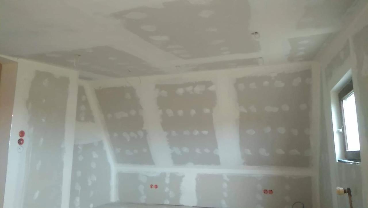 Pokój na poddaszu w trakcie wykańczania płytami gipsowo-kartonowymi, widoczne szpachlowane łączenia i otwory na gniazdka elektryczne, okno w prawym górnym rogu.