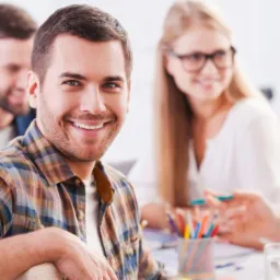 Uśmiechnięty mężczyzna w kraciastej koszuli siedzi przy stole z grupą osób, w tle kolorowe kredki w pojemniku i rozmyte postacie w trakcie dyskusji.