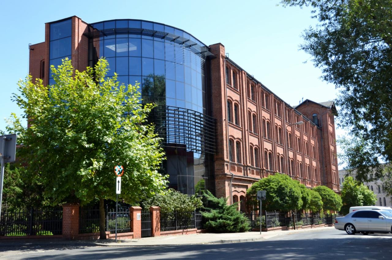 Budynek szkoły językowej z czerwonej cegły z nowoczesną, przeszkloną dobudówką, widoczny od strony ulicy z drzewami i ogrodzeniem.