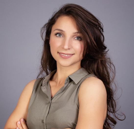 Portret uśmiechniętej kobiety z brązowymi włosami i zielonymi oczami na szarym tle, ubrana w oliwkową koszulę bez rękawów, z założonymi rękami.