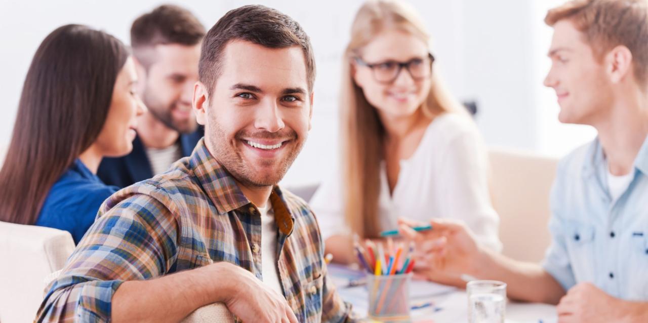 Uśmiechnięty mężczyzna w kraciastej koszuli siedzi przy stole z grupą osób, w tle kolorowe kredki w pojemniku i rozmyte postacie w trakcie dyskusji.