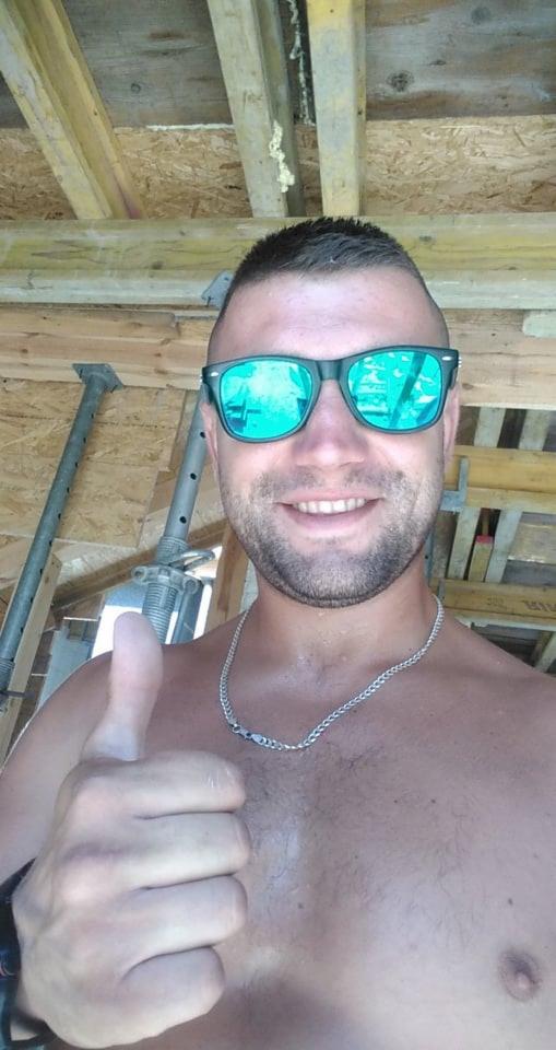 Uśmiechnięty mężczyzna w okularach przeciwsłonecznych, dający kciuk w górę, na tle drewnianej konstrukcji stropu w trakcie budowy.
