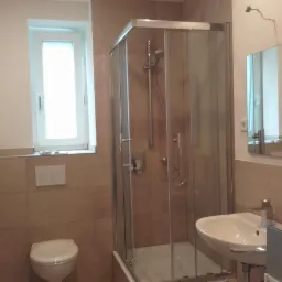 Wykończona łazienka z kabiną prysznicową, umywalką, grzejnikiem drabinkowym i lustrem, wykończona beżowymi płytkami ceramicznymi.