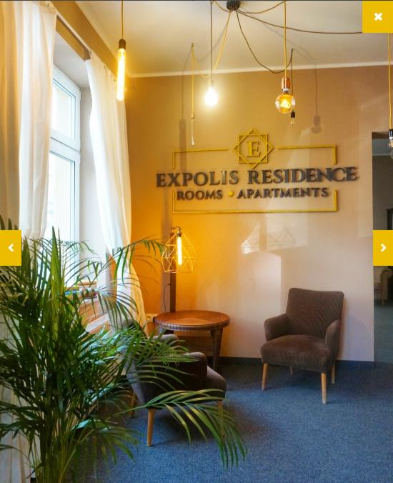 Wnętrze apartamentu z dwoma fotelami, okrągłym stolikiem i rośliną doniczkową, oświetlone wiszącymi lampami z żarówkami Edisona, na ścianie logo Expolis Residence Rooms Apartments.