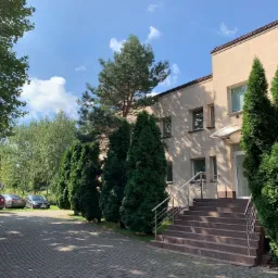 Dwupiętrowy budynek biurowy z beżową elewacją, otoczony zielonymi drzewami i krzewami, z parkingiem dla samochodów na brukowanym podjeździe.