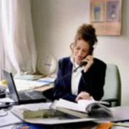 Kobieta w biurowym stroju rozmawia przez telefon, przeglądając dokumenty na biurku z laptopem i zegarem w tle, oświetlona naturalnym światłem z okna.