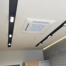Biały sufit z centralnie umieszczonym kwadratowym panelem klimatyzacji, oświetlenie punktowe w czarnych listwach, fragment ściany z szarej cegły i jasne meble w tle.