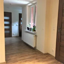 Korytarz z podłogą wyłożoną panelami imitującymi drewno, białym grzejnikiem pod oknem i drewnianymi drzwiami z przeszkleniami w tle.