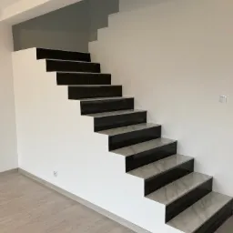 Nowoczesne schody z czarno-szarymi płytkami imitującymi marmur, osadzone na białej konstrukcji, w minimalistycznym wnętrzu z drewnopodobną podłogą.