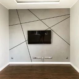 Szara ściana z dekoracyjnymi czarnymi liniami tworzącymi abstrakcyjny wzór, podwieszony telewizor, ciemna drewniana podłoga i białe listwy przypodłogowe.