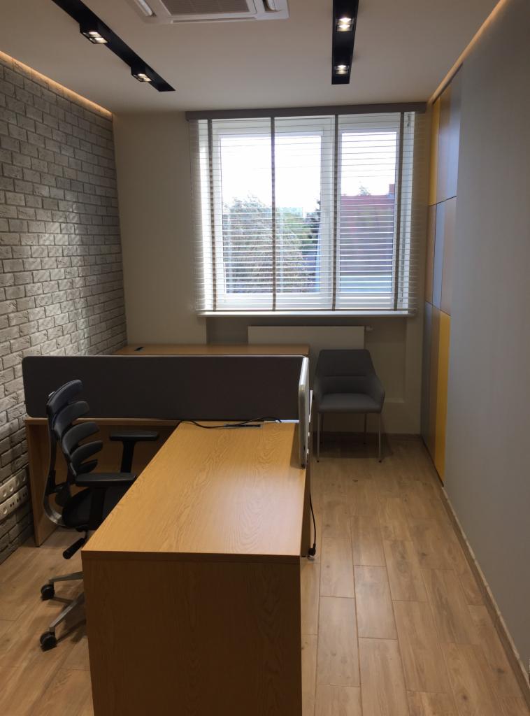 Nowoczesne biuro w Sierpcu z drewnianym biurkiem, szarą ścianą z cegły i oknem z żaluzjami, oświetlone listwami LED.