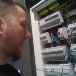 Mężczyzna z brodą, w czarnej koszulce z logo, pochyla się nad białą skrzynką rozdzielczą z bezpiecznikami i przewodami, podłączając niebieski kabel.