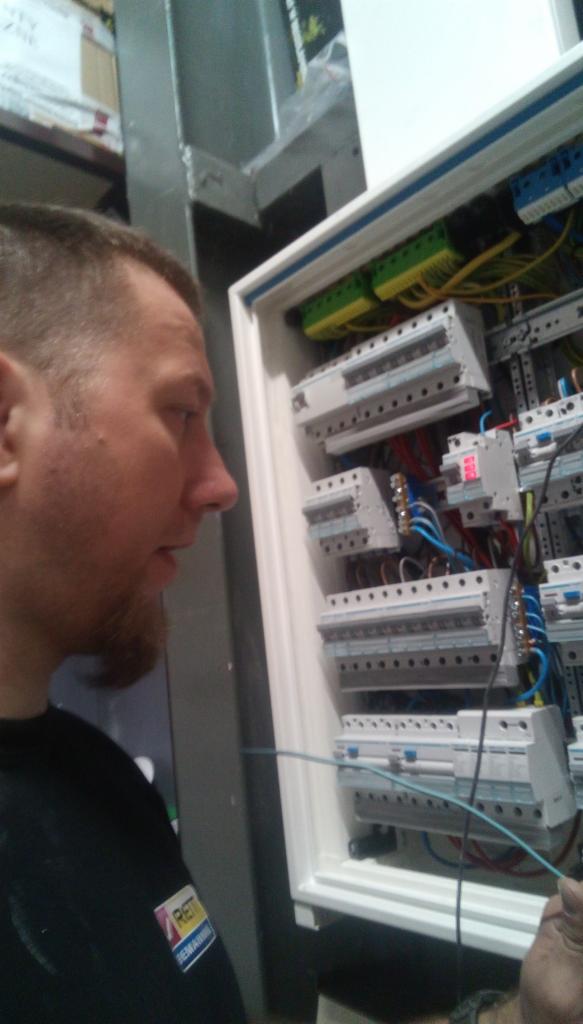 Mężczyzna z brodą, w czarnej koszulce z logo, pochyla się nad białą skrzynką rozdzielczą z bezpiecznikami i przewodami, podłączając niebieski kabel.