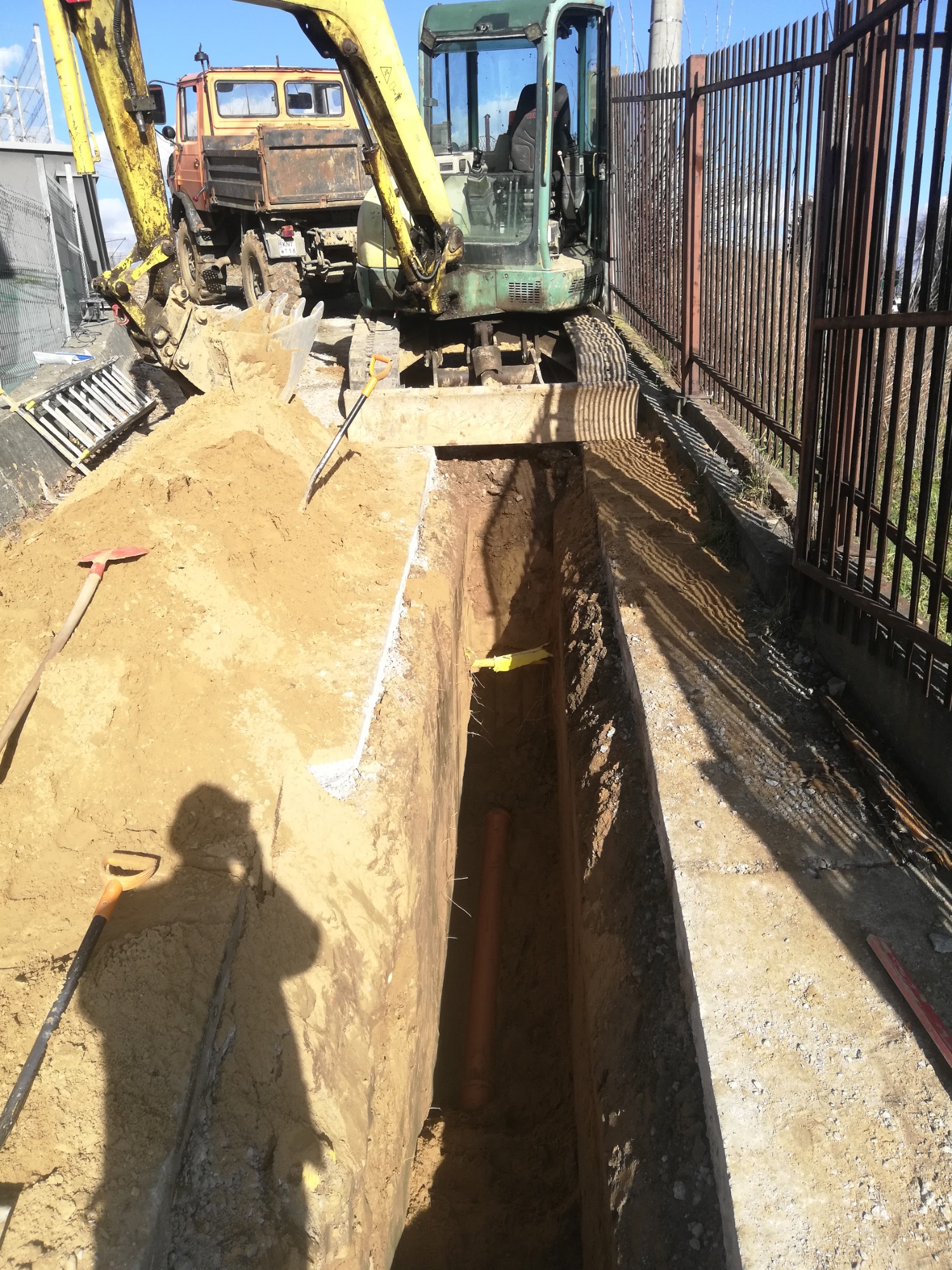 Wąski wykop w ziemi z pomarańczową rurą na dnie, obok zaparkowany stary, pomarańczowy samochód ciężarowy i mała koparka, cień osoby na skarpie wykopu.