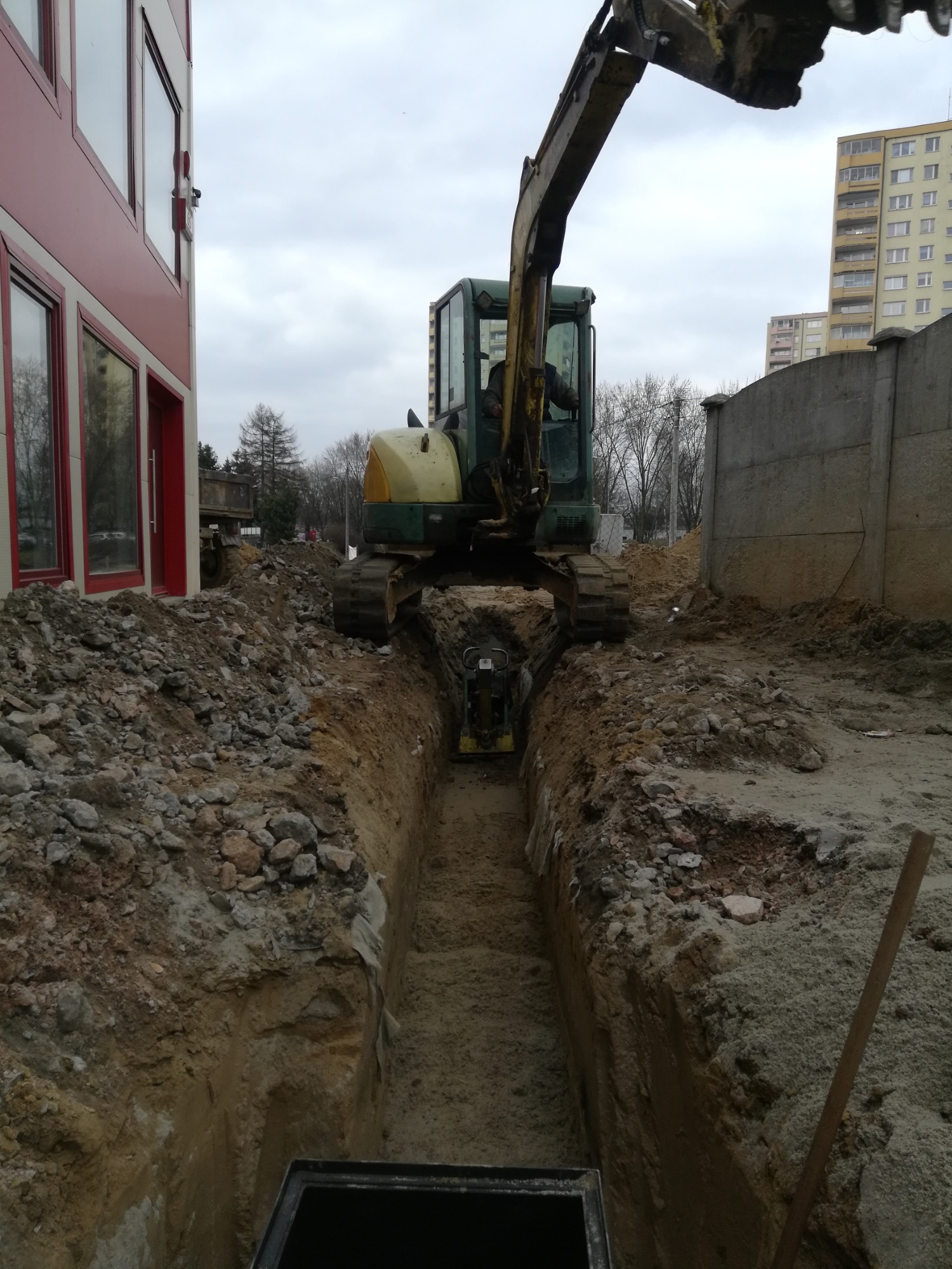 Żółto-zielona koparka gąsienicowa ustawiona nad wąskim wykopem, w tle budynek z czerwonymi ramami okiennymi i blok mieszkalny.