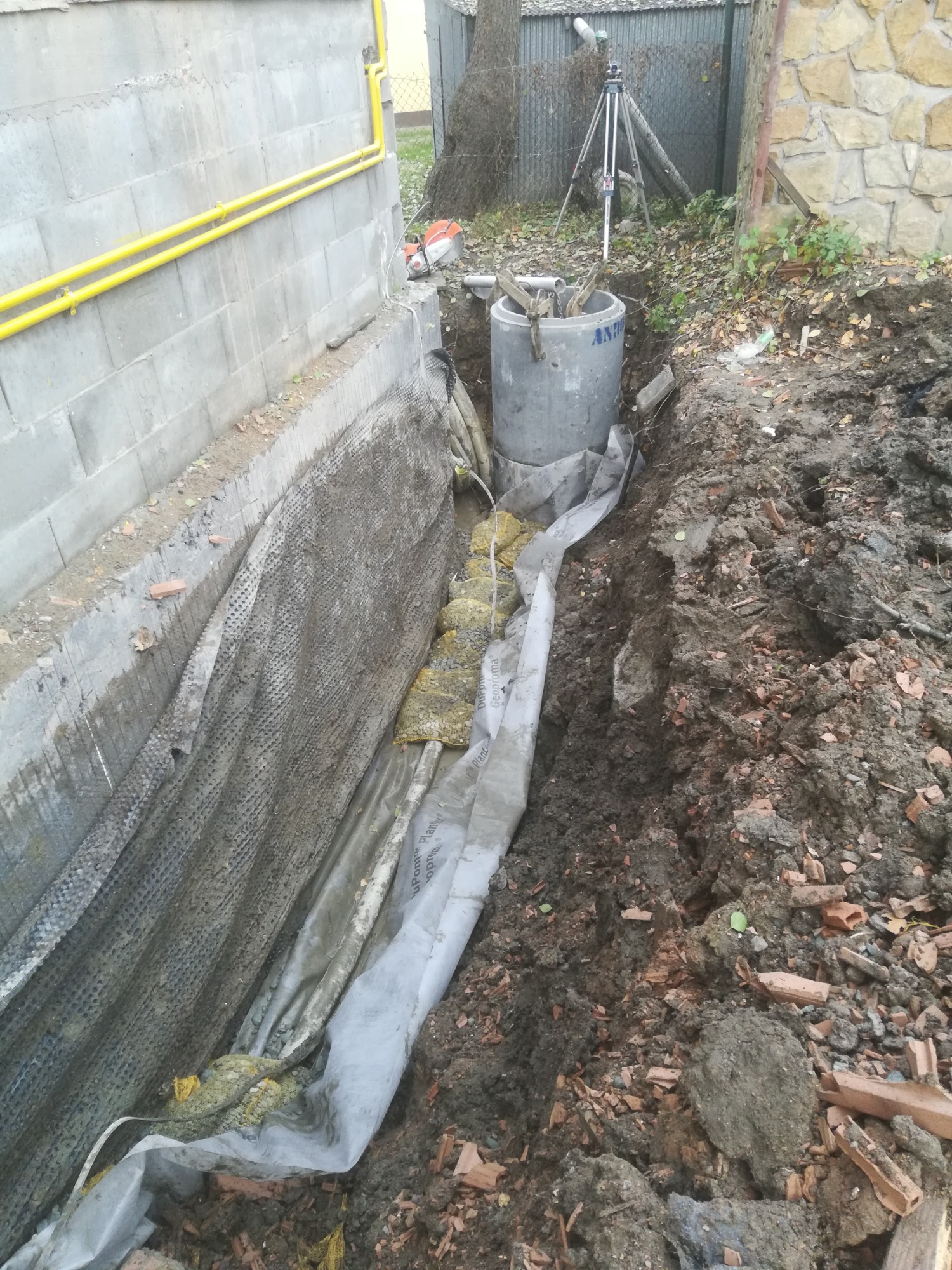 Betonowy krąg studni umieszczony w wykopie, zabezpieczony geowłókniną i żółtą izolacją, widoczne rury przy budynku i niwelator w tle.