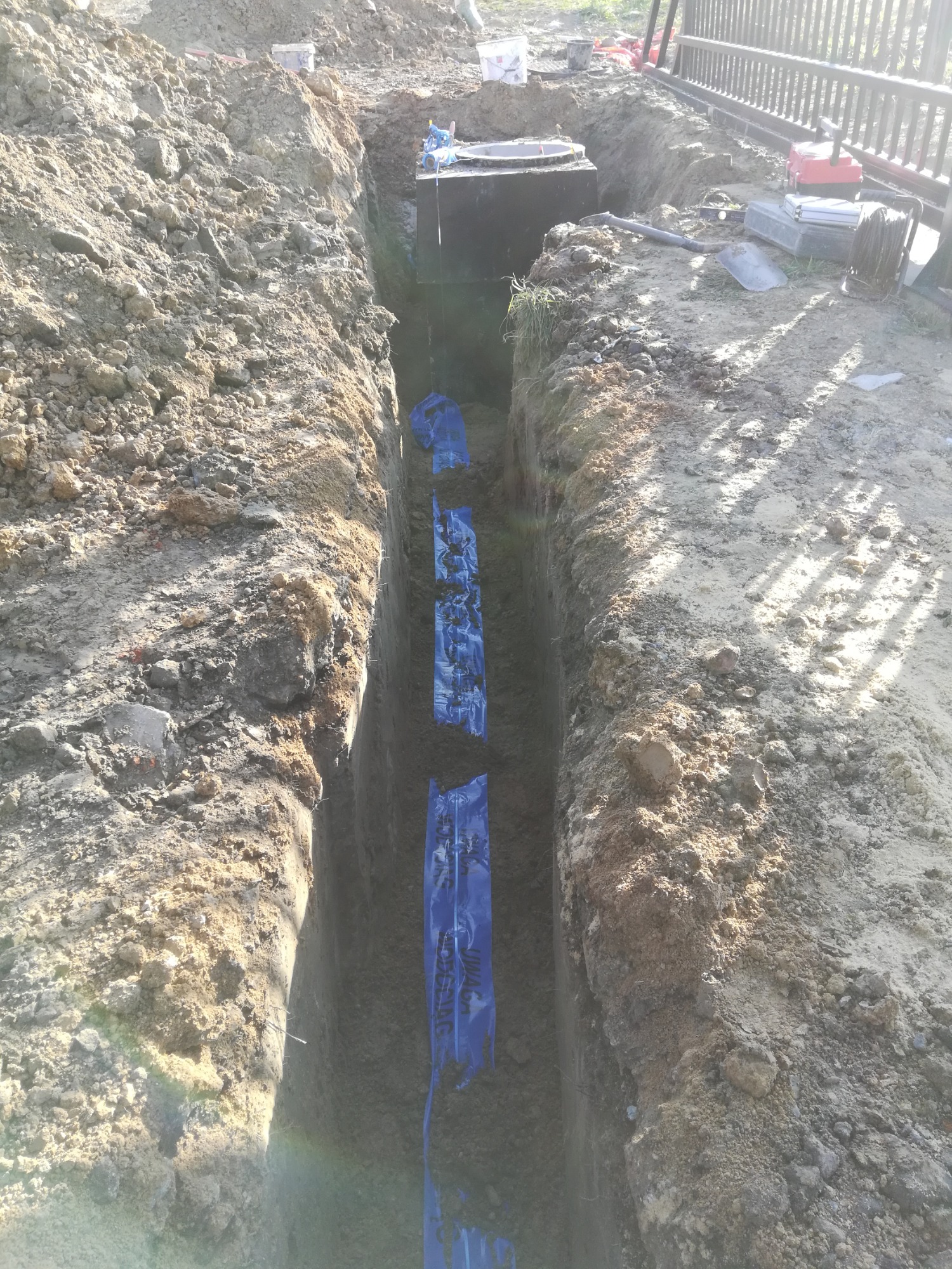 Długi, wąski wykop w ziemi z niebieską folią zabezpieczającą, widoczny fragment studzienki kanalizacyjnej z pokrywą na końcu wykopu, narzędzia i akcesoria budowlane na brzegu wykopu.