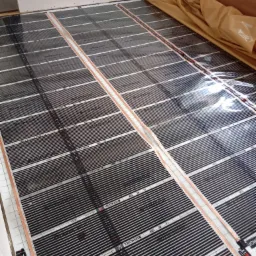 Rozłożona folia grzewcza Termofol na podłodze, przygotowana do montażu ogrzewania podłogowego, widoczne równe pasy z czarnymi elementami grzewczymi i białymi przerwami.