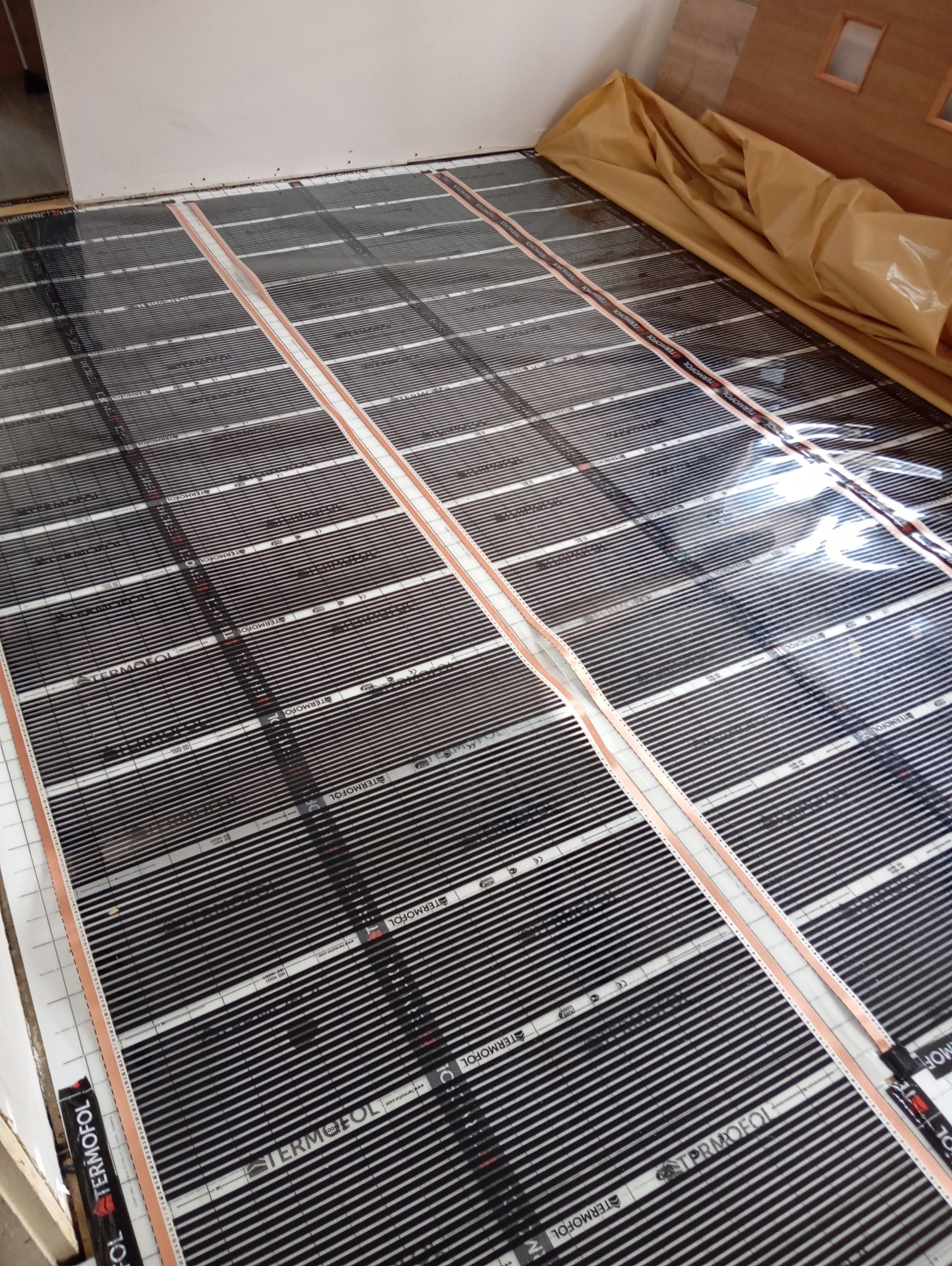 Rozłożona folia grzewcza Termofol na podłodze, przygotowana do montażu ogrzewania podłogowego, widoczne równe pasy z czarnymi elementami grzewczymi i białymi przerwami.