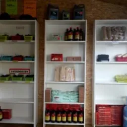 Wnętrze składu opału z trzema białymi regałami wypełnionymi różnorodnym towarem, w tym brykietem, płynami, drobnymi akcesoriami i materiałami budowlanymi.