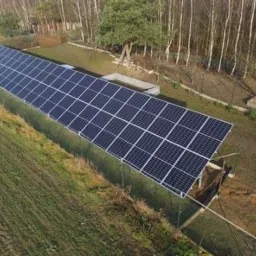 Długa instalacja fotowoltaiczna na gruncie, otoczona siatką, widok z góry, w tle las.