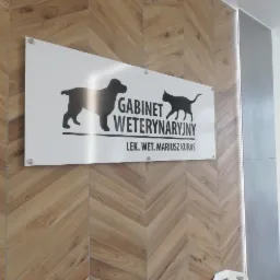Tablica informacyjna Gabinetu Weterynaryjnego z sylwetkami psa i kota, przymocowana do ściany wyłożonej drewnopodobną okładziną w jodełkę.