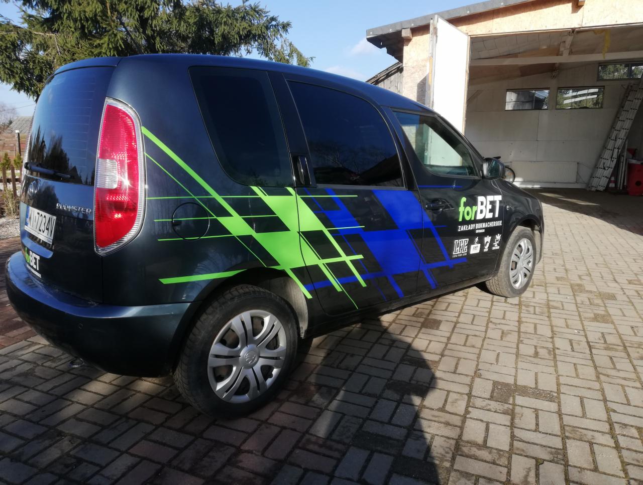 Ciemny samochód dostawczy Skoda Roomster z zielono-niebieską grafiką geometryczną i logo 'forBET zakłady bukmacherskie' na boku, zaparkowany na brukowanym podjeździe przed garażem.