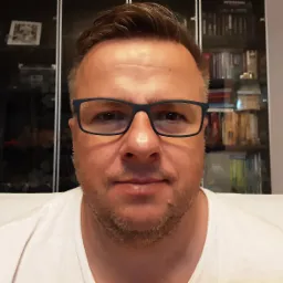 Portret mężczyzny w okularach na tle regału z książkami i pamiątkami, prawdopodobnie właściciel firmy przeprowadzkowej.