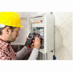 Mężczyzna w kasku mierzy parametry w rozdzielnicy elektrycznej za pomocą multimetru cyfrowego. Widoczne bezpieczniki, przewody i elementy instalacji.