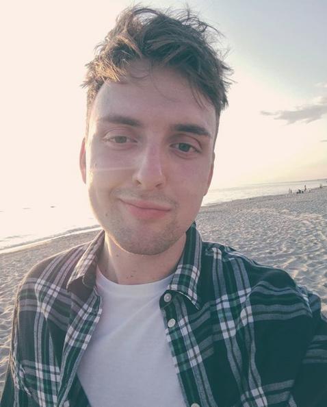 Młody mężczyzna w koszuli w kratę robi selfie na plaży o zachodzie słońca. W tle widać morze i piasek. Oświetlenie słoneczne tworzy ciepłą atmosferę.