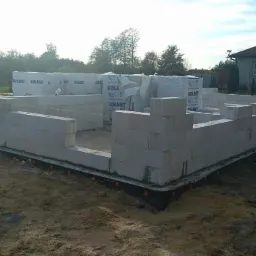 Wznoszenie ścian fundamentowych z bloczków betonowych Solbet na czarnej hydroizolacji z pomarańczowymi wypustkami, w tle zapakowane bloczki i dom jednorodzinny.