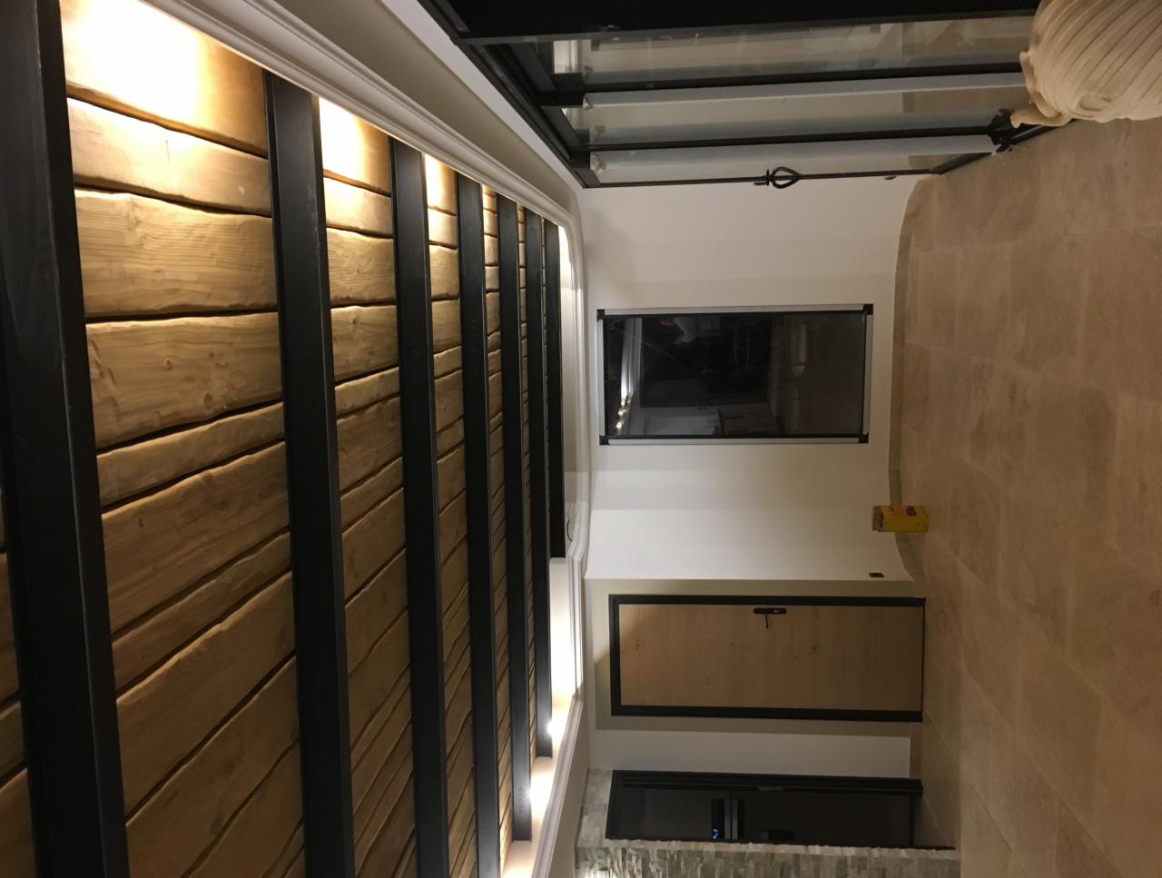 Drewniana ściana z czarnymi, pionowymi elementami dekoracyjnymi i podświetleniem, widok na fragment pomieszczenia z oknem i beżową podłogą.