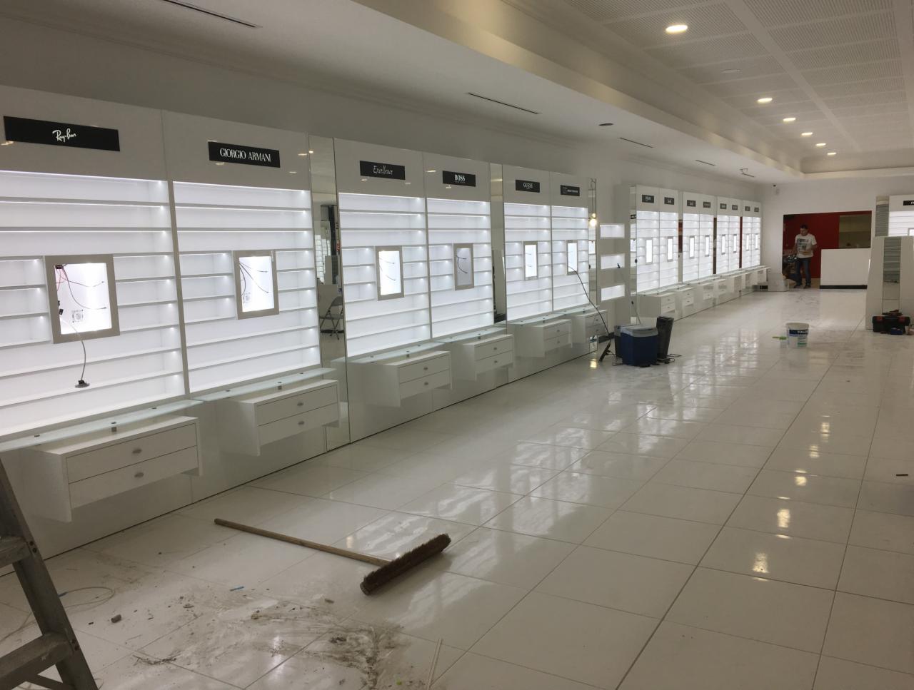 Wnętrze sklepu optycznego w trakcie wykańczania: białe, podświetlane regały z szufladami pod markami Ray-Ban, Giorgio Armani, Excellence, Boss i Guess, biała podłoga w kwadratowe płytki, widoczny...