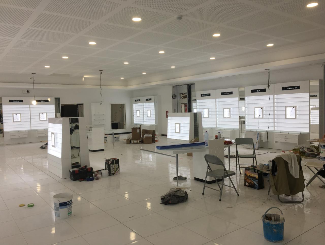 Wnętrze sklepu optycznego w trakcie prac wykończeniowych: białe, podświetlane ekspozytory na okulary, rozstawione krzesła, puszki z farbą i narzędzia na błyszczącej podłodze z płytek, sufit...