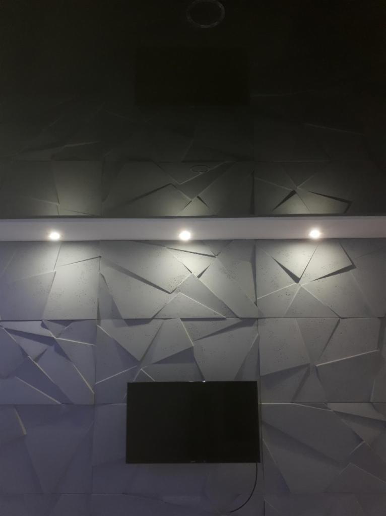 Szara ściana z trójwymiarową, geometryczną fakturą oświetlona przez trzy punktowe lampy, z zamontowanym telewizorem na dole.