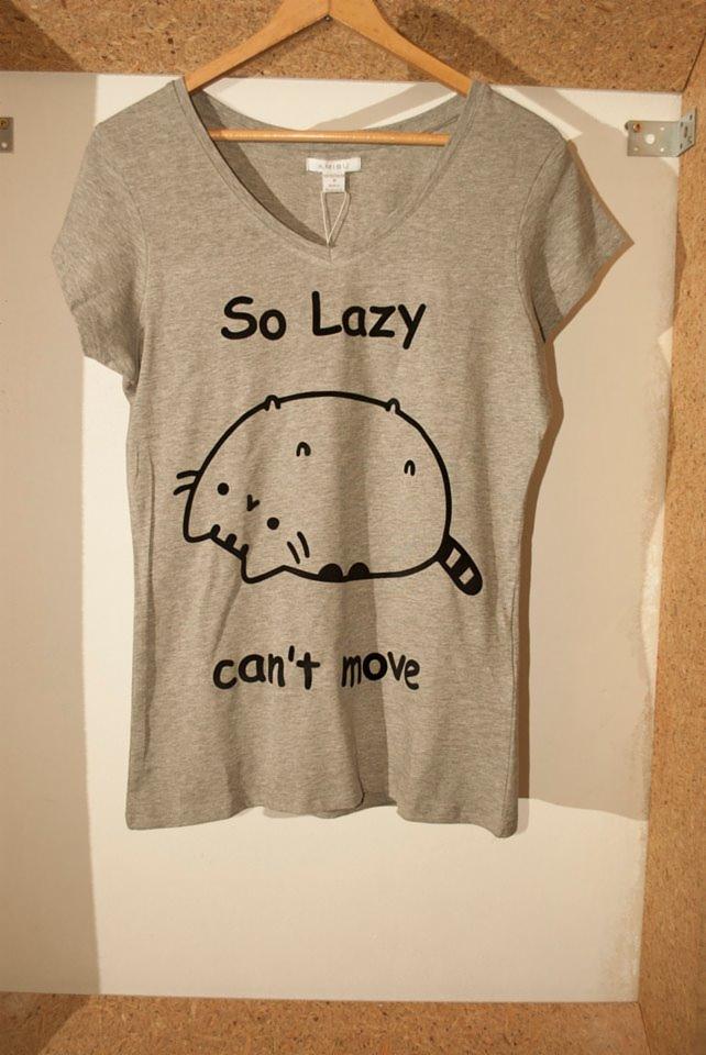 Szara koszulka z nadrukiem leniwego kota i napisem 'So Lazy can't move', wisząca na wieszaku na tle korkowej ściany.