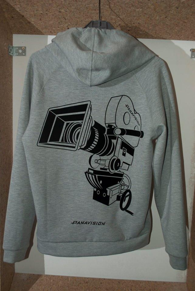 Szara bluza z kapturem z czarnym nadrukiem kamery filmowej Panavision, wisząca na wieszaku na tle korkowej ściany.