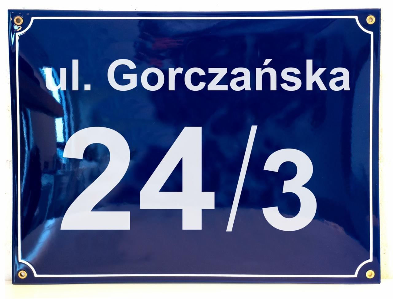 Emaliowana tabliczka z adresem 'ul. Gorczańska 24/3' w kolorze granatowym z białymi napisami, przymocowana na rogach za pomocą złotych nitów.