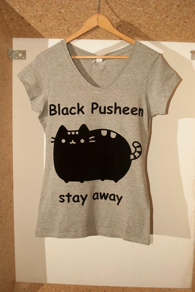 Szara koszulka z nadrukiem czarnego kota Pusheen z napisem 'Black Pusheen stay away', wisząca na drewnianym wieszaku na tle ściany.