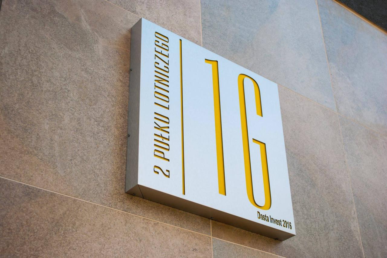 Tablica informacyjna z nazwą ulicy i logo, wykonana z metalu z żółtymi akcentami, zamontowana na szarej kamiennej ścianie.