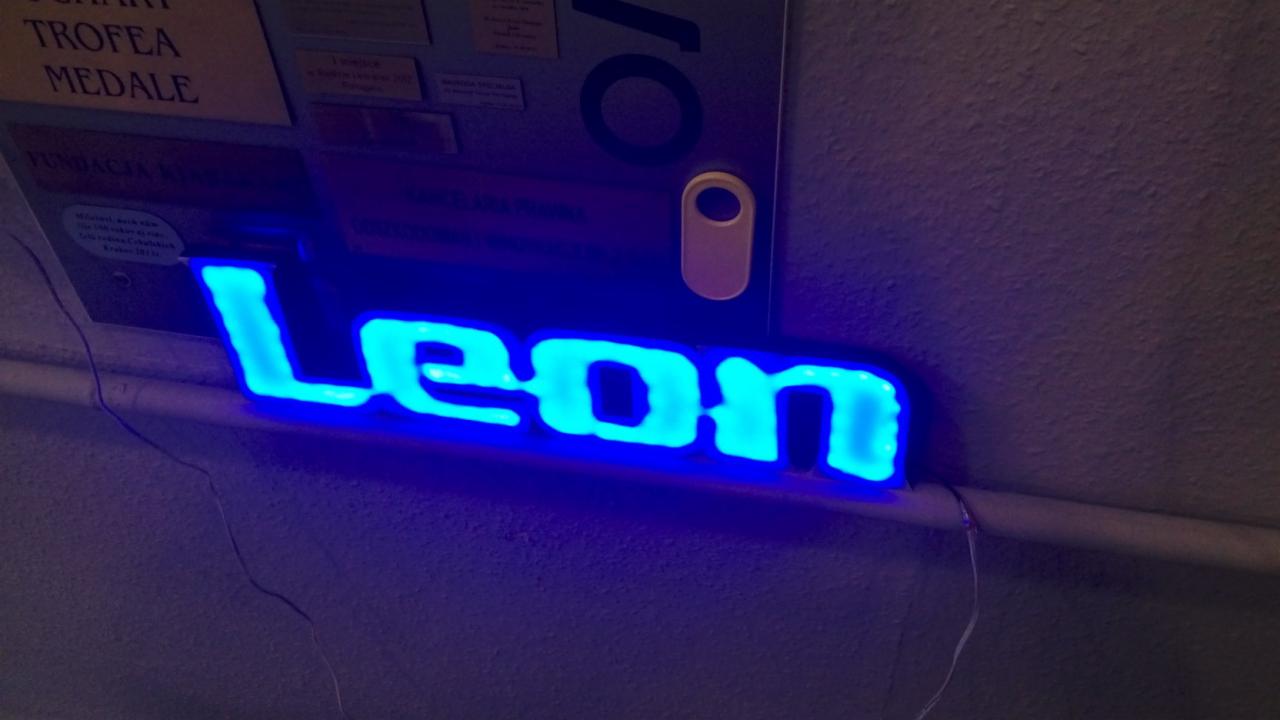 Podświetlany niebieski napis 'Leon' zamontowany na ścianie, widoczne kable zasilające.