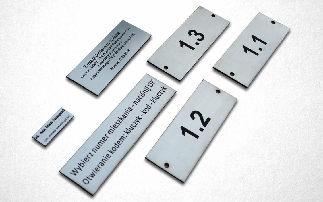 Metalowe tabliczki z grawerowanymi napisami i numerami, w tym tabliczka upamiętniająca 50-lecie Instytutu Katalizy i Fizykochemii Powierzchni PAN oraz tabliczki z numerami 1.1, 1.2, 1.3...