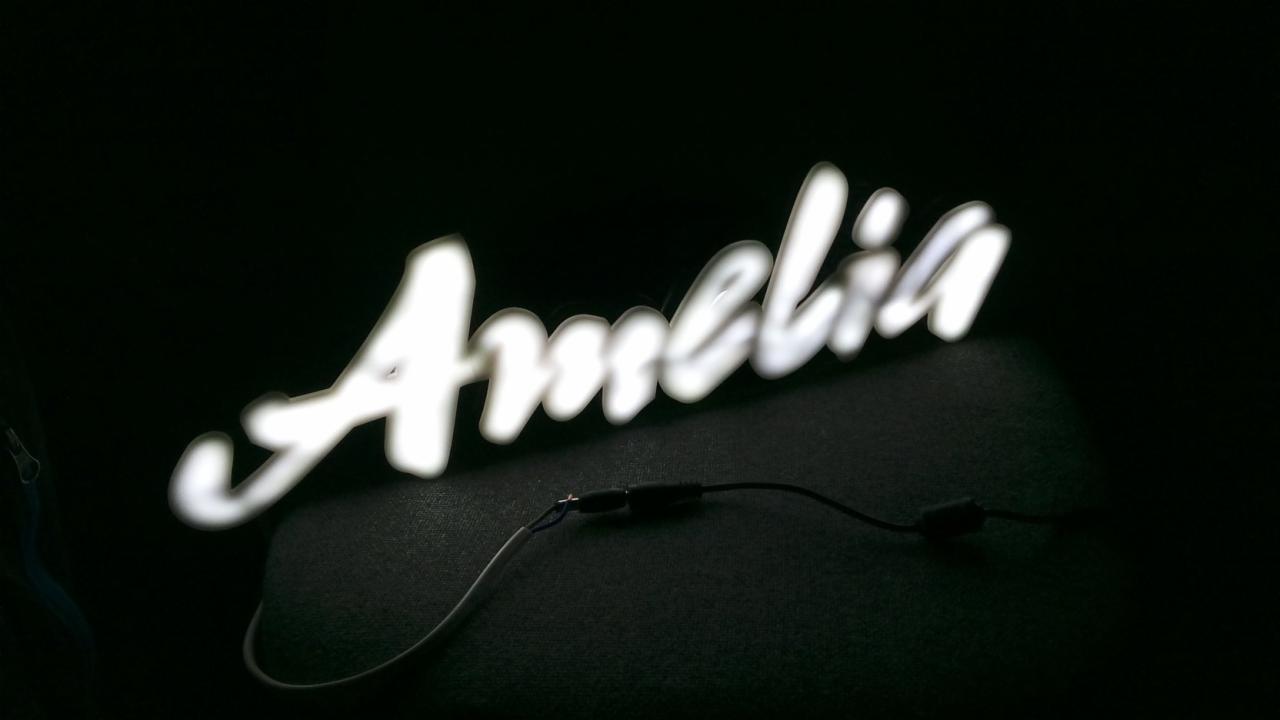 Świecący, ręcznie pisany napis 'Amelia' z podłączonymi kablami na ciemnym tle.