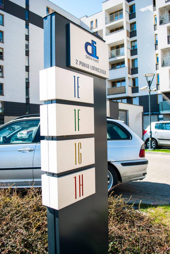 Oznakowanie budynku mieszkalnego z logo 'dasta invest' i numerami mieszkań w kolorach niebieskim, zielonym, złotym i czerwonym, na tle nowoczesnej architektury.
