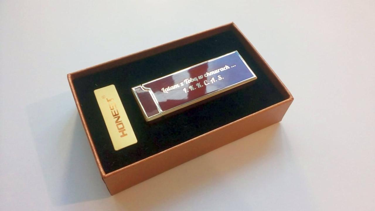 Zapalniczka marki Honest w brązowym pudełku z czarnym wnętrzem, z wygrawerowanym tekstem 'Latam z Tobą w chmurach... 1.K.K.C.A.S.', obok złoty element z napisem Honest.