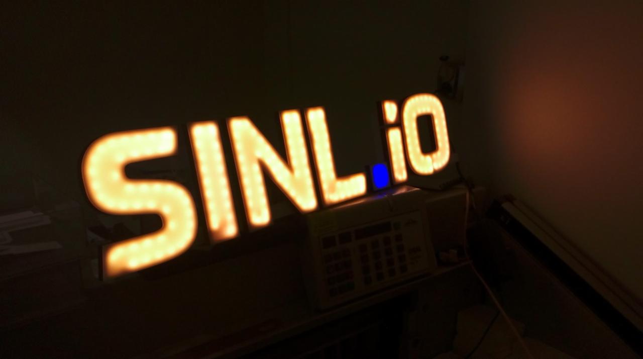 Świecący, przestrzenny napis SINLio z podświetleniem w ciepłym odcieniu żółci, widoczny na tle ściany i częściowo na tle biurka z kablem i kalkulatorem.