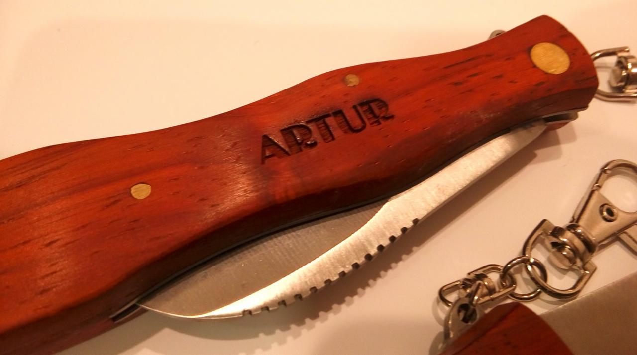 Otwarty scyzoryk z drewnianą rękojeścią z wygrawerowanym napisem 'ARTUR' oraz brelokiem, wykonany w Krakowie.