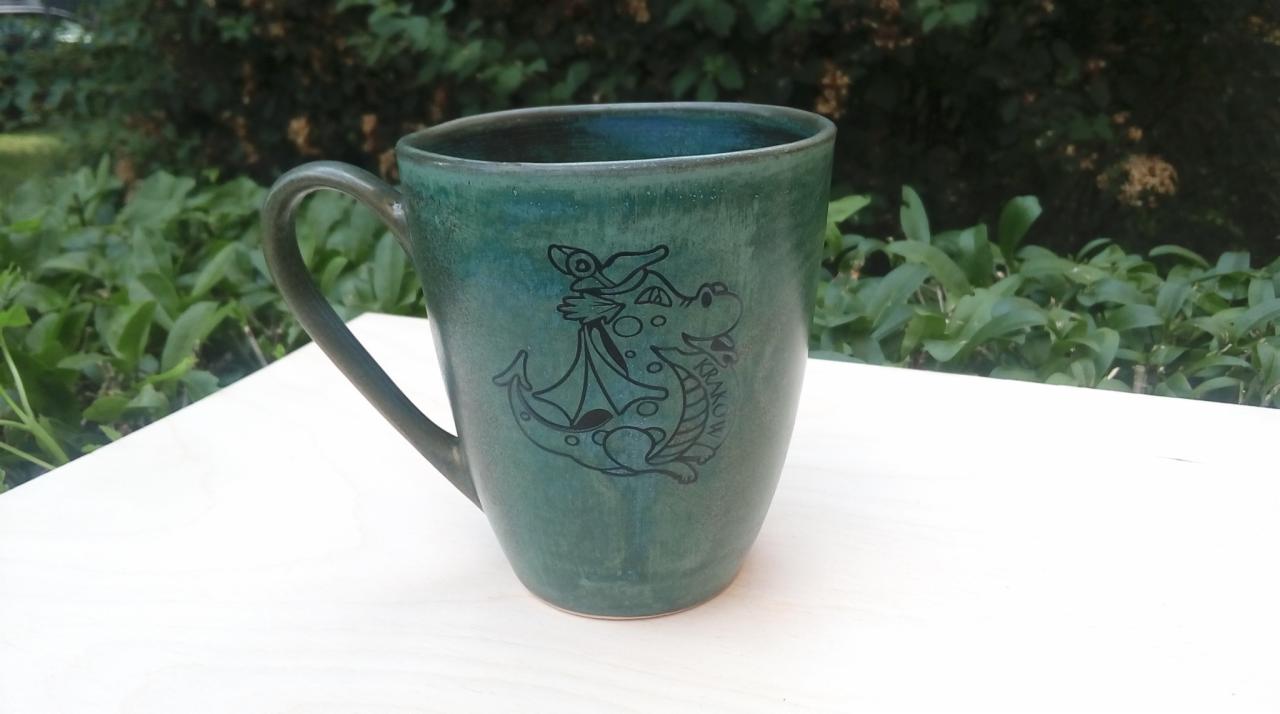Zielony, ceramiczny kubek z czarnym rysunkiem smoka wawelskiego i napisem 'Kraków' na jasnym blacie na tle zieleni.