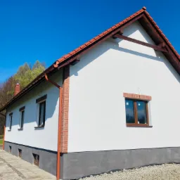 Świeżo otynkowany, biały dom jednorodzinny z czerwoną dachówką i ceglanymi narożnikami, widok z boku w słoneczny dzień. Widoczne okna z brązowymi ramami i szara podmurówka. Na ziemi rozsypany żwir.